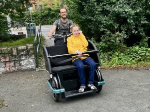 Eine Frau mit gelber Jacke sitzt vorne in einer Fahrradrikscha. Ein Mann dahinter fährt diese Rikscha