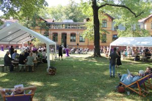 eine Wiese vor einem großen Haus. Auf Stühlen und Bänken, auf Zelten und im freien sitzen Menschen