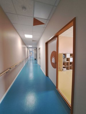 ein Flur mit blauem Boden. Links ein Handlauf. Rechts eine Tür mit beigem Rahmen. Auf der Tür ist ein Piktogramm abgebildet. an der Decke zeigt ein Pfeil in der gleichen Farbe zum Raum hin.