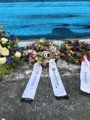 vor einer blauen Wand liegt ein kleiner Trauerkranz. Die Aufschrift auf der Schleife: Wir erinnern! und das Logo der leben lernen gGmbH