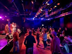 Auf einer B&uuml;hne (links) spielt eine Band. Das Publikum tanzt.