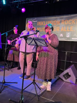 Ein Mann und eine Frau stehen auf einer B&uuml;hne und singen in ein Mikrophon. Im Hintergrund steht eine Frau , die Gitarre spielt