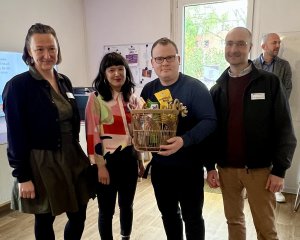 Vier Menschen gucken in die Kamera. Einer h&auml;lt einen Geschenkkorb im Arm